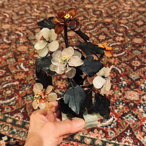 May include: A decorative floral sculpture featuring translucent flowers in shades of white, amber, and orange, with yellow centers and red accents. The flowers are attached to dark green leaves and stems, mounted on a rectangular base.