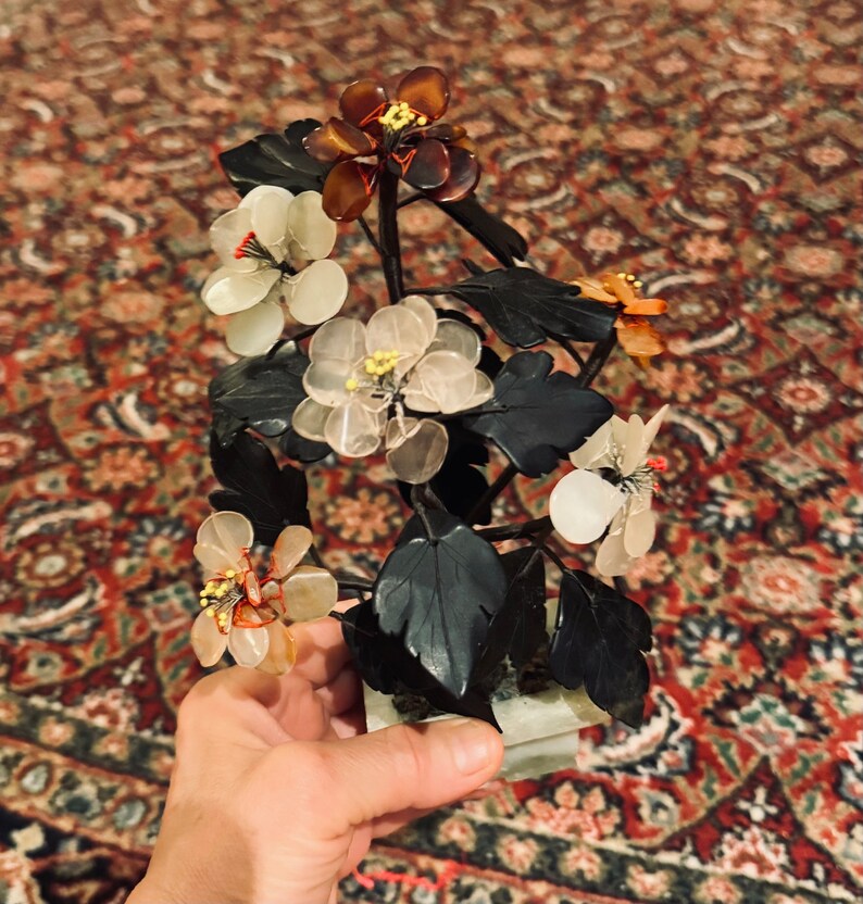 May include: A decorative floral sculpture featuring translucent flowers in shades of white, amber, and orange, with yellow centers and red accents. The flowers are attached to dark green leaves and stems, mounted on a rectangular base.