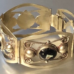 Puede incluir: Una pulsera articulada plateada con dise&ntilde;os calados decorativos y detalles de piedras preciosas negras. La pulsera presenta una serie de paneles rectangulares unidos por bisagras, con detalles ornamentados y piedras negras circulares.