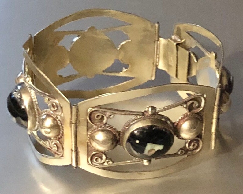 Puede incluir: Una pulsera articulada plateada con dise&ntilde;os calados decorativos y detalles de piedras preciosas negras. La pulsera presenta una serie de paneles rectangulares unidos por bisagras, con detalles ornamentados y piedras negras circulares.