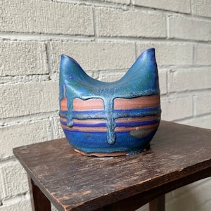 Op de afbeelding: Een blauwe en bruine keramische kattenfiguur met een druppelglazuur. Het beeldje heeft een rond lichaam en twee puntige oren.