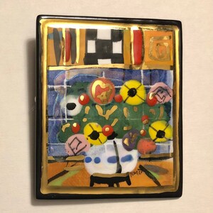May include: A colorful ceramic tile with a black frame and gold accents. The tile features a stylized floral arrangement with red, yellow, and blue flowers. The tile is signed "Tomie" in the lower right corner.