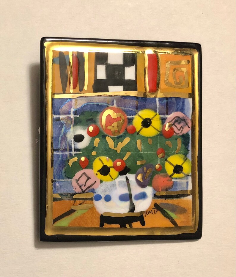 May include: A colorful ceramic tile with a black frame and gold accents. The tile features a stylized floral arrangement with red, yellow, and blue flowers. The tile is signed "Tomie" in the lower right corner.
