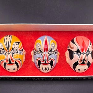 Puede incluir: Tres máscaras decorativas de ópera china en una caja rectangular con interior rojo. Las máscaras presentan diseños intrincados en amarillo, azul, rojo y negro, mostrando el arte facial tradicional.