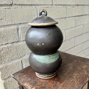 Peut inclure: Un pot en céramique noire avec un couvercle. Le pot est recouvert d'un glaçage vert et a la forme d'une double gourde. Le pot est posé sur une table en bois.