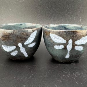 May include: Two dark blue ceramic bowls with white dragonfly designs. The bowls are slightly different shades of blue and have a textured surface.
