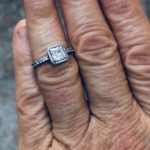 May include: A silver ring with a square-shaped clear stone set in a halo of smaller stones.