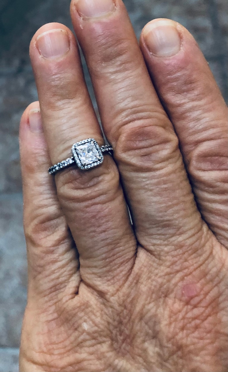 May include: A silver ring with a square-shaped clear stone set in a halo of smaller stones.