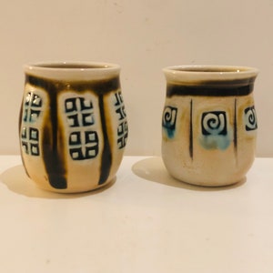 May include: Two small ceramic mugs with brown and white geometric designs. One mug has a blue and white swirl design, while the other has a black and white geometric design.