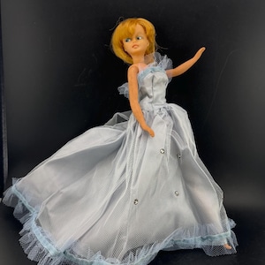 Puede incluir: Muñeca vintage con cabello rubio vestida con un vestido azul claro. El vestido presenta una capa transparente con adornos delicados y un dobladillo con volantes. La muñeca está en movimiento, con un brazo levantado. El fondo es negro liso.