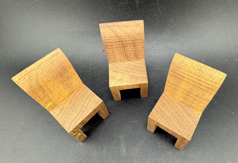 May include: Three small, wooden chairs with a natural wood grain finish. Each chair has a curved back and a rectangular base with a small, black opening. The chairs are arranged on a dark surface.