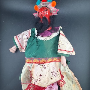 May include: A doll with a bright red face, black beard, and a silver headpiece adorned with colorful pom-poms. The doll wears a green tunic, a patterned cream-colored apron with geometric shapes, and green boots, featuring intricate fabric textures.