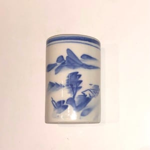 May include: A cylindrical, white ceramic container with a blue landscape design. The container features a rounded top and is adorned with hand-painted blue mountain and tree motifs, creating a serene, artistic aesthetic. The container is likely for storage or display.