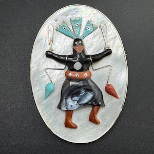 May include: An oval brooch featuring a central inlay figure of a dancer crafted from black stone, orange stone, and mother-of-pearl. The figure holds two silver-toned swords and wears a headdress of three turquoise pieces against a mother-of-pearl background.