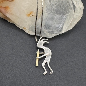 May include: A silver-tone pendant necklace featuring a stylized Kokopelli figure holding a flute. The pendant has a gold-colored flute and hangs from a delicate silver chain. The necklace is displayed against a dark gray background.
