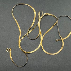 May include: A collection of gold-colored herringbone chain necklaces. One necklace is fully extended, while the others are partially coiled. The necklaces have a clasp closure. The background is a dark gray color.