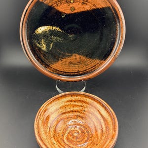 May include: Two handmade ceramic pieces: a large plate and a small bowl. The plate has a dark brown rim and a black centre with gold accents. The bowl has a spiral design in shades of brown and orange.