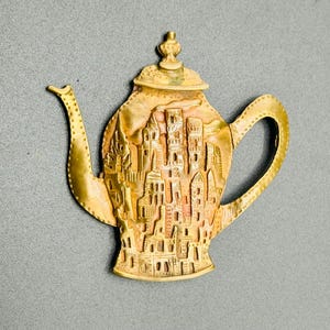 May include: A gold-colored teapot with a detailed design of a city on the front. The teapot has a small lid and a spout.
