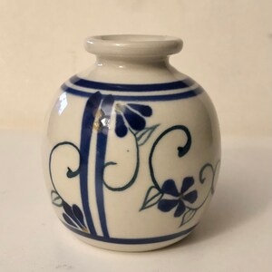 May include: A small, white ceramic vase with blue and gold floral designs. The vase has a rounded body and a narrow neck.