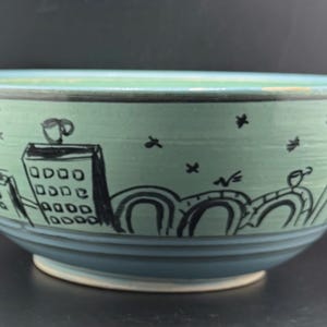 May include: A ceramic bowl with a light teal exterior and a blue interior. The bowl is decorated with a black cityscape design, including buildings, bridges, and stars. A coffee cup is also depicted.