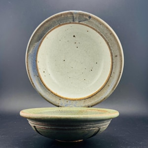 May include: Two ceramic bowls. The top bowl has a light blue and brown rim with a speckled interior. The bottom bowl is green with a brown rim and horizontal grooves. The bowls are set against a dark background.