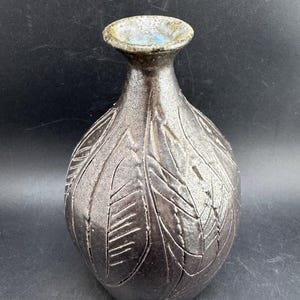 Può includere: Un vaso in ceramica grigio metallizzato scuro con corpo bulboso e collo stretto. La superficie presenta un motivo a foglie intagliato e testurizzato su tutto l'esterno. Il bordo del vaso ha una forma leggermente svasata con smalto interno azzurro.