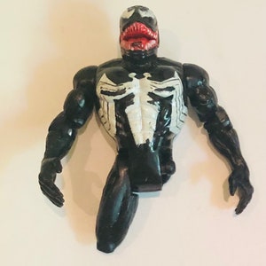 May include: Black and white plastic action figure of Venom, a Marvel Comics superhero. The figure has a large mouth with red teeth and a white symbol on its chest.