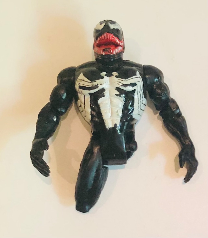 May include: Black and white plastic action figure of Venom, a Marvel Comics superhero. The figure has a large mouth with red teeth and a white symbol on its chest.