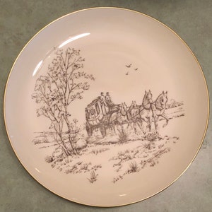 May include: A vintage pink and gold ceramic plate with a brown line drawing of a horse-drawn carriage and trees. The plate has a gold rim.