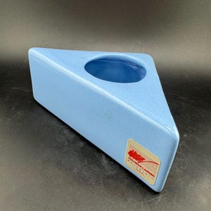 May include: A light blue, triangular ceramic vase with a circular opening. The vase has a matte finish and a small label on the side. The label reads "Peltman Langer Porcelain USA".