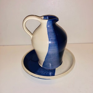 Puede incluir: Una jarra de cerámica blanca y azul con asa y un platillo a juego. La jarra tiene una franja azul en el lateral.