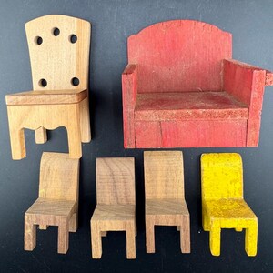 May include: A collection of miniature wooden furniture pieces, including a light-colored chair with a perforated back, a red armchair, and several small chairs in shades of brown and yellow. The pieces are arranged on a dark surface.