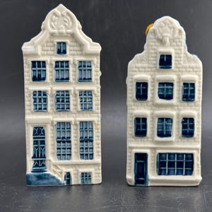 Könnte beinhalten: Zwei keramische Miniatur-Kanalhaus-Figuren mit weißer Basis und blau bemalten Details. Jedes Bauwerk zeigt ein Treppengiebel-Dach, mehrere rechteckige Fenster und eine Eingangstür, die der Architektur traditioneller niederländischer Häuser nachempfunden sind.