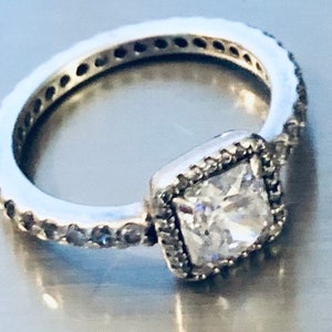 May include: A silver ring with a square-shaped clear stone set in a bezel setting. The ring is surrounded by a row of smaller clear stones.