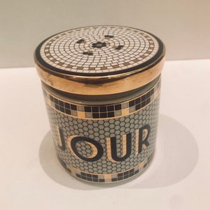 May include: A white ceramic jar with a gold lid and black and gold mosaic design. The jar has the word "JOUR" printed on the front in black letters.