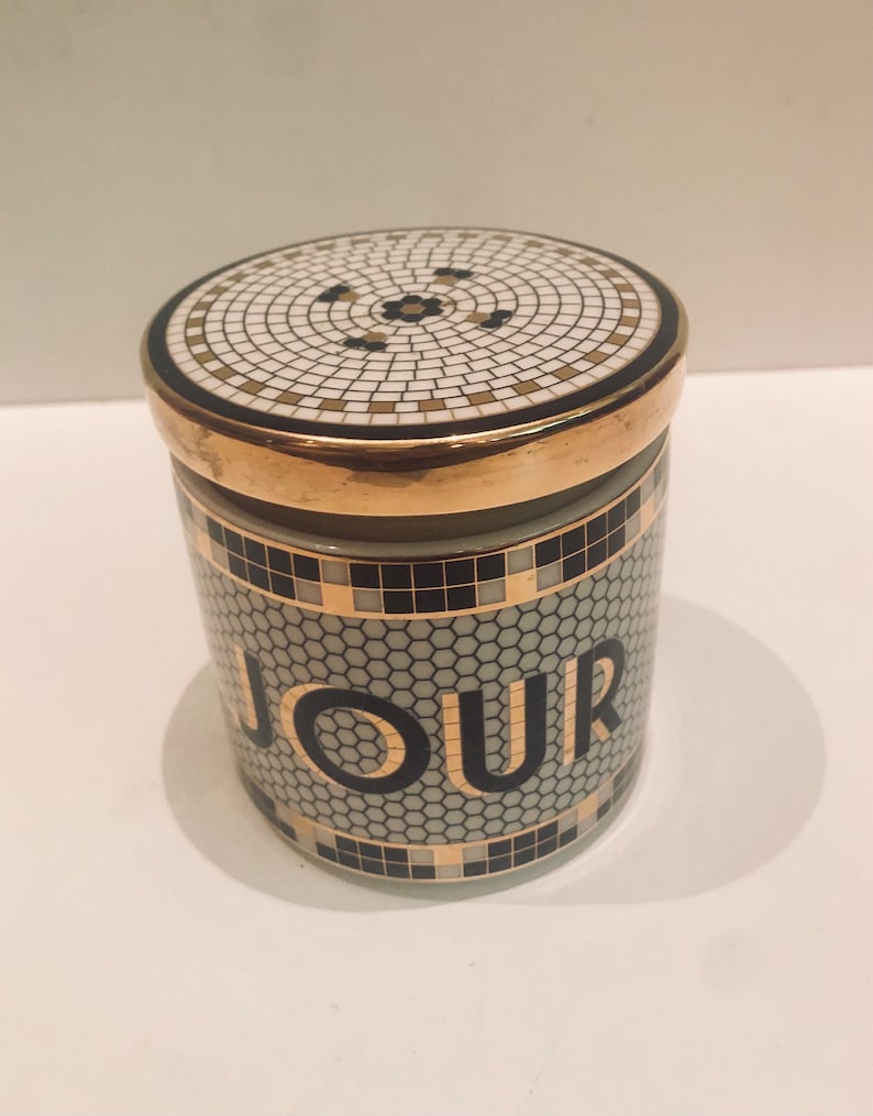 May include: A white ceramic jar with a gold lid and black and gold mosaic design. The jar has the word "JOUR" printed on the front in black letters.