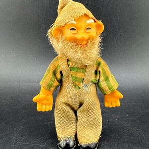 May include: A vintage gnome doll with a smiling face, a long, tan beard, and a pointed hat. The doll wears a green and yellow striped shirt, brown overalls, and black shoes. The doll is approximately 6 inches tall.
