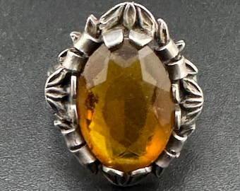 May include: An oval-shaped, silver-toned brooch with a large, faceted amber-colored stone at its center. The stone is surrounded by a decorative silver setting with floral and ribbon-like details. The brooch is set against a dark background.