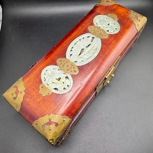 May include: A rectangular wooden box with a reddish-brown finish, adorned with brass corner accents and decorative jade-like carvings. The box features an oval jade carving in the center and smaller carvings on the top.