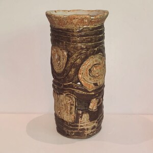 May include: A brown ceramic vase with a textured surface and a white glaze. The vase has a cylindrical shape and is decorated with a pattern of circles and swirls.