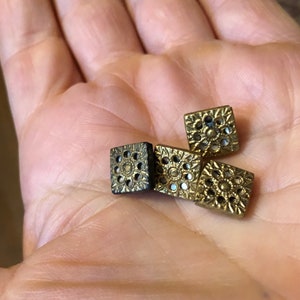 May include: Four gold-coloured square buttons with intricate designs. The buttons have a raised, circular pattern with small holes throughout.