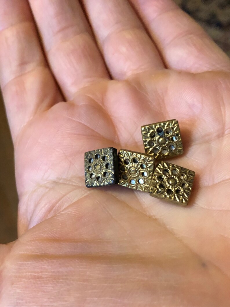 May include: Four gold-coloured square buttons with intricate designs. The buttons have a raised, circular pattern with small holes throughout.