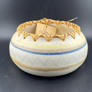 Puede incluir: Un cuenco redondo de cerámica blanquecina con rayas horizontales azules, amarillas y marrones. El borde está adornado con paja tejida y una etiqueta cuelga del borde. El cuenco presenta un patrón geométrico.