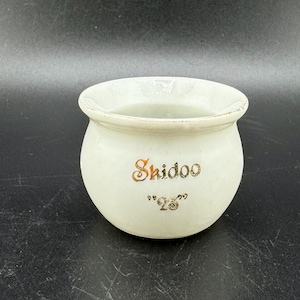 Könnte beinhalten: Weißer Keramiktopf mit einer goldenen Inschrift, die "Skidoo '28" lautet.