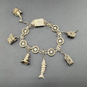 May include: Silver-toned charm bracelet featuring various detailed charms. The bracelet includes charms shaped like a pagoda, fish, birdcage, and other decorative elements. The bracelet is displayed on a dark gray surface.
