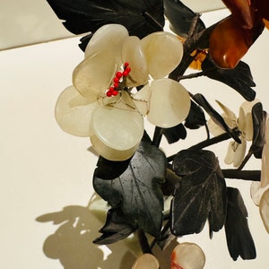 May include: A decorative floral sculpture featuring clusters of pale, translucent stone grapes and dark green leaves. Small red beads accent the grapes. The sculpture is set against a white background, creating a striking contrast.