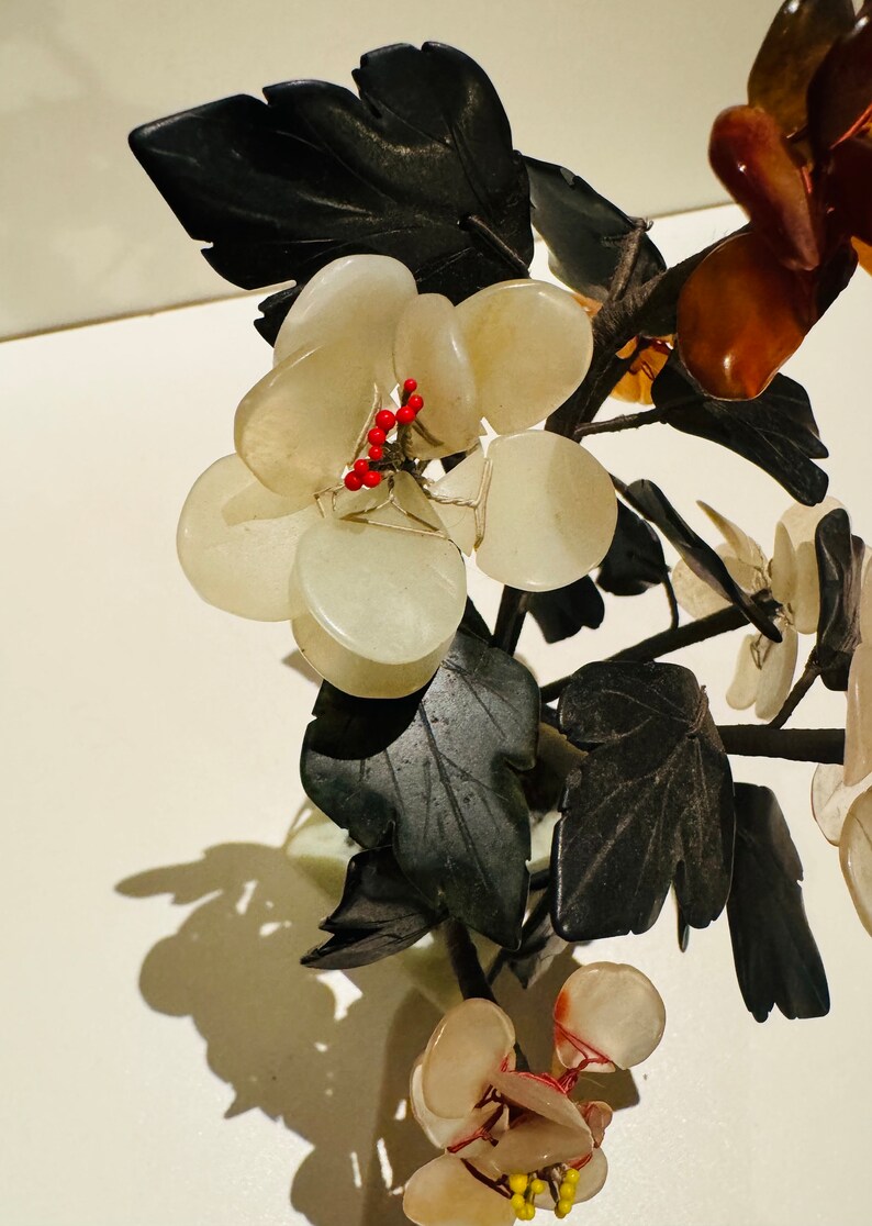 May include: A decorative floral sculpture featuring clusters of pale, translucent stone grapes and dark green leaves. Small red beads accent the grapes. The sculpture is set against a white background, creating a striking contrast.