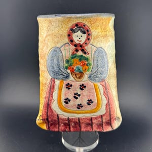 Puede incluir: Un jarrón rectangular de cerámica con un diseño de arte popular. El jarrón representa a una mujer con vestimenta tradicional sosteniendo una cesta de flores. La mujer lleva un pañuelo rojo con lunares negros y un delantal a rayas rojas y blancas. El jarrón está sostenido por un soporte transparente.