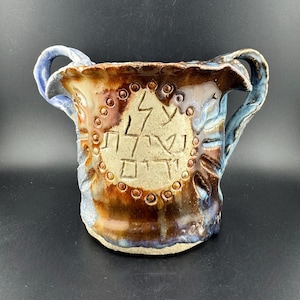 Könnte beinhalten: Eine braune Keramikvase mit zwei Henkeln und einem kreisförmigen Design mit hebräischen Buchstaben. Die Vase hat eine blaue Glasur und braune Akzente.