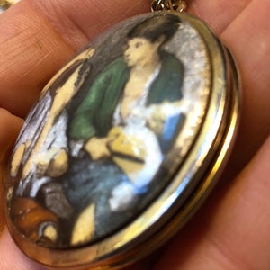 May include: Oval pendant necklace with a gold-tone chain. The pendant features a miniature painting of a person wearing a green jacket and white shirt. The painting has a gold-tone border.